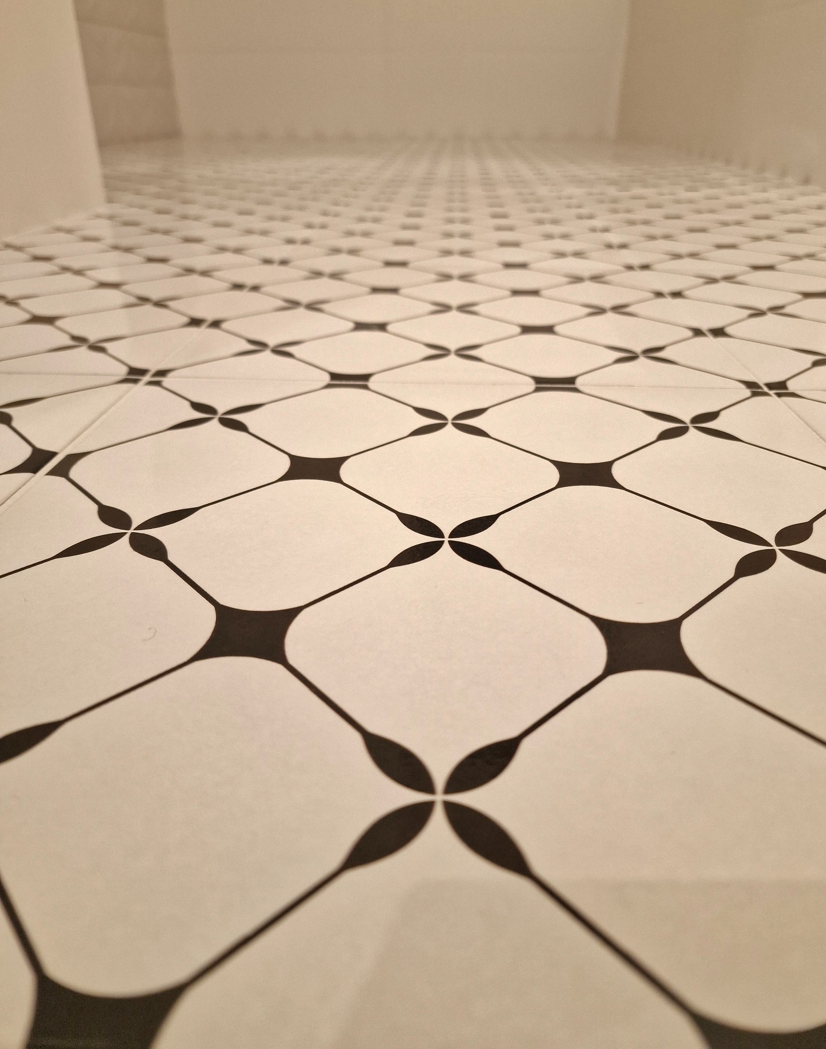 Białe płytki podłogowe z czarnym wzorem w kształcie kwiatów, ułożone w łazience. Perspektywa z dołu, widoczny fragment ściany w tle.