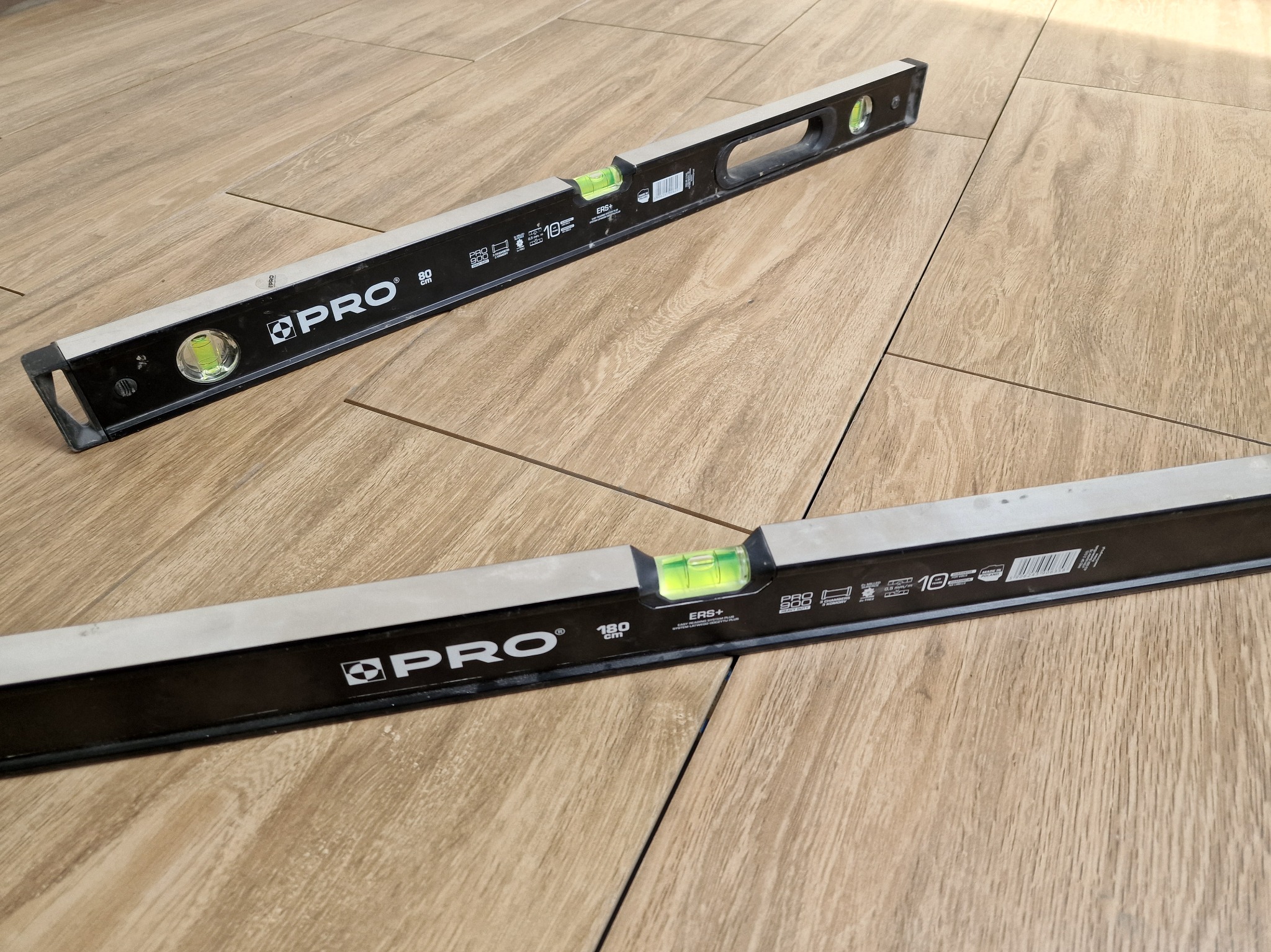 Ułożone płytki podłogowe imitujące drewno, sprawdzane poziomicą. Widoczne dwie poziomice marki PRO, leżące na jasnych płytkach z wyraźnym wzorem drewna.