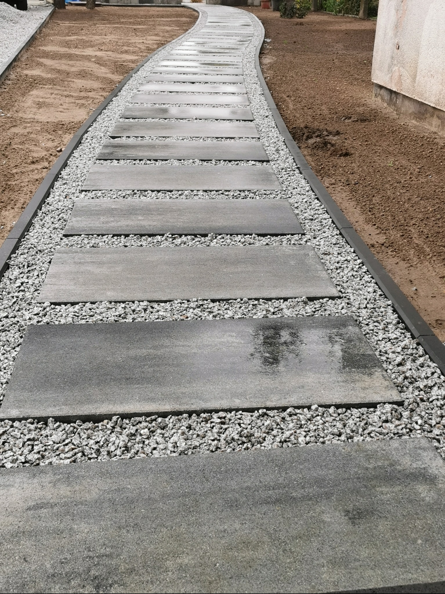 Ścieżka z szarych płyt betonowych, wysypana drobnym, jasnym żwirem, obrzeża z ciemnego tworzywa. Widok z perspektywy, lekko zakrzywiona trasa.