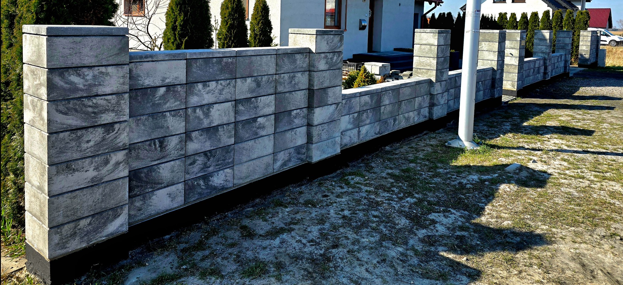 Nowoczesny, niski murek z szarych, betonowych bloczków o marmurkowym wzorze, oddzielający posesję. Widoczne słupki i podmurówka. Tło stanowi dom i tuje.