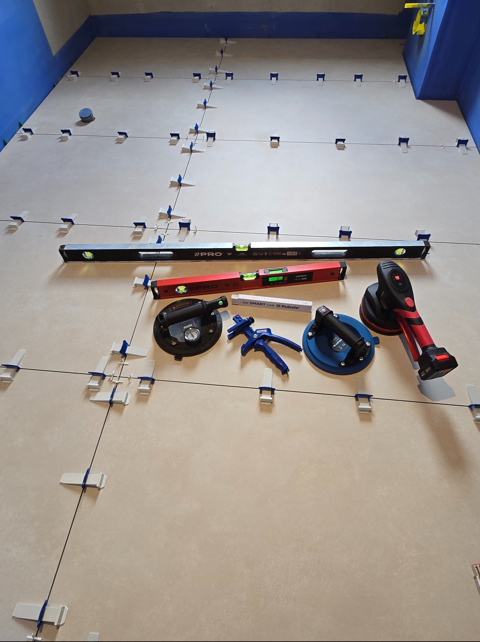 Ułożone płytki podłogowe z systemem poziomowania, poziomice, przyssawki do płytek i narzędzia glazurnicze w trakcie pracy. Widok z góry na podłogę w niebieskim pomieszczeniu.