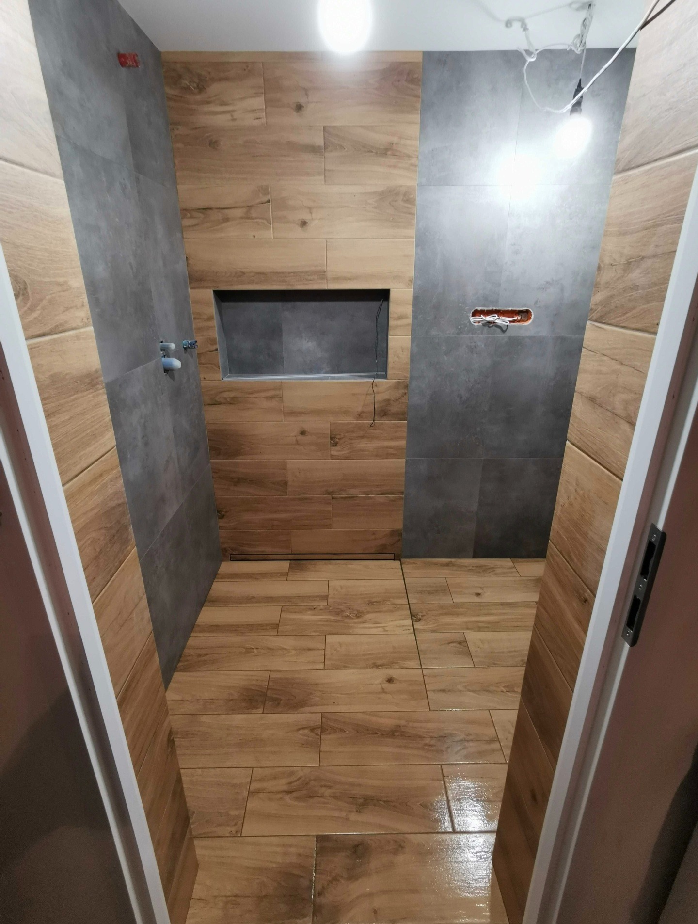 Wykończona łazienka z drewnopodobnymi i szarymi płytkami, wnęką w ścianie i widoczną instalacją elektryczną. Widok zza otwartych drzwi.