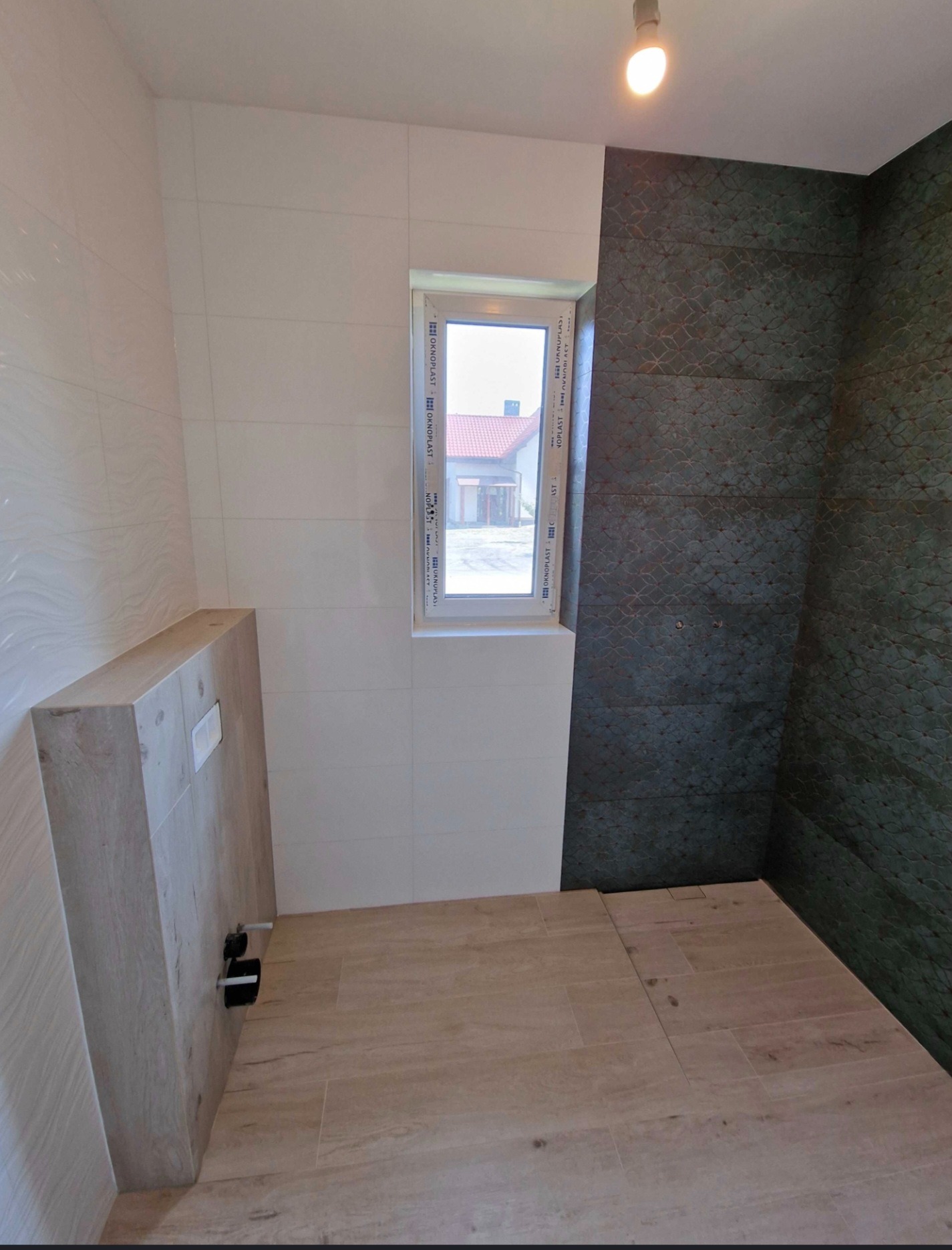 Nowoczesna łazienka po remoncie: beżowe płytki podłogowe i imitujące drewno obudowy WC, białe i grafitowe płytki ścienne, okno z widokiem na dom.