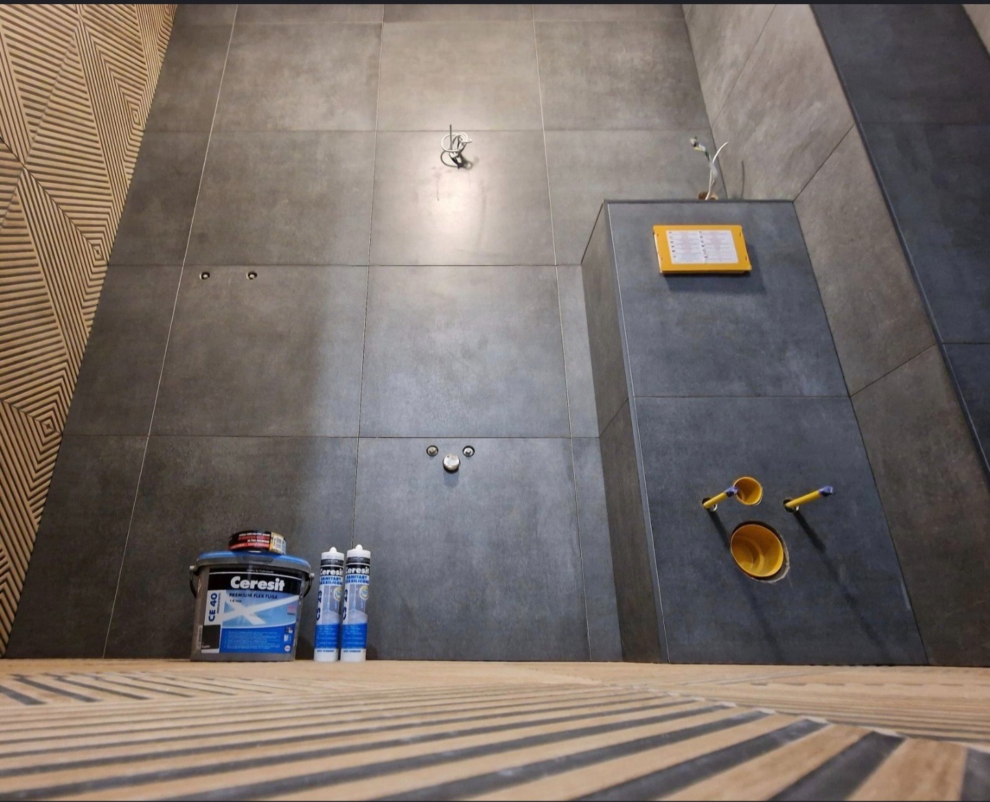 Nowoczesna łazienka w szarościach z akcentem drewna. Widoczne płytki, instalacja podtynkowa i materiały budowlane. Ujęcie z dołu podkreśla wysokość pomieszczenia.