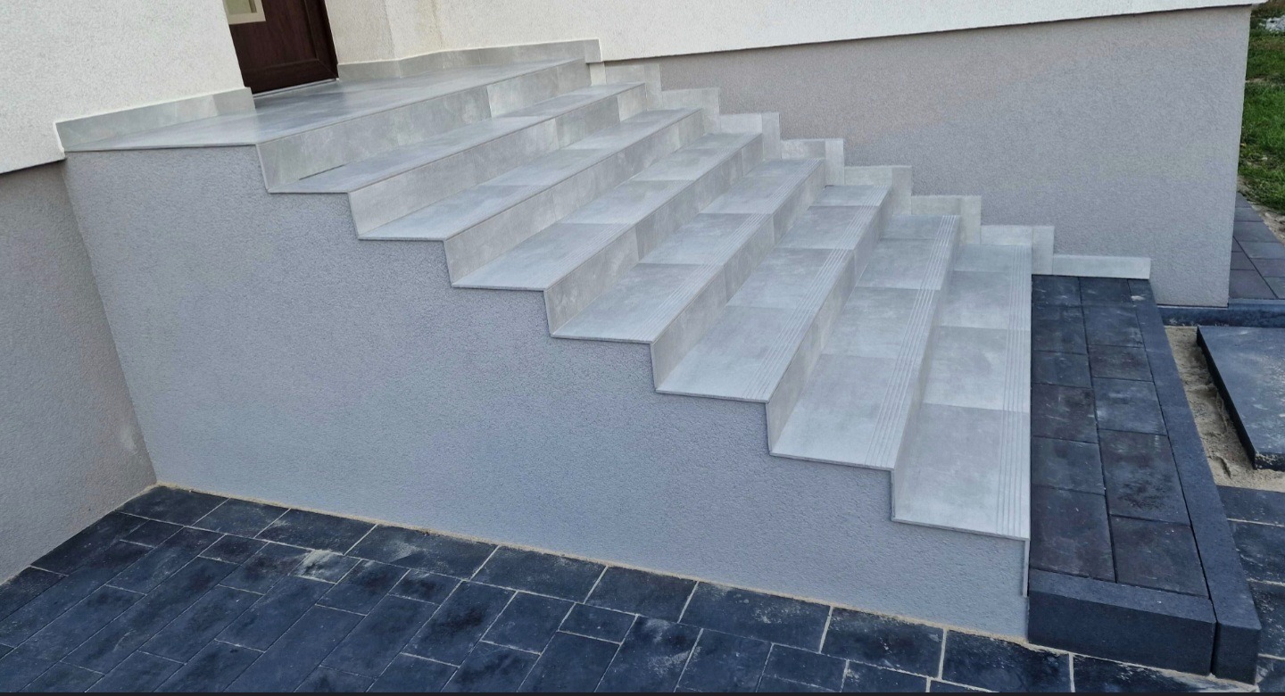 Zewnętrzne schody wyłożone szarymi płytkami, z subtelnymi rowkami antypoślizgowymi. Ciemna kostka brukowa u podstawy i obok schodów. Elewacja budynku w odcieniach szarości.