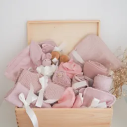 Drewniana skrzynia wypełniona różową wyprawką dla niemowlaka: kocyki w kropki, ręczniki, ubranka i pluszowy króliczek. Całość przewiązana białymi wstążkami.