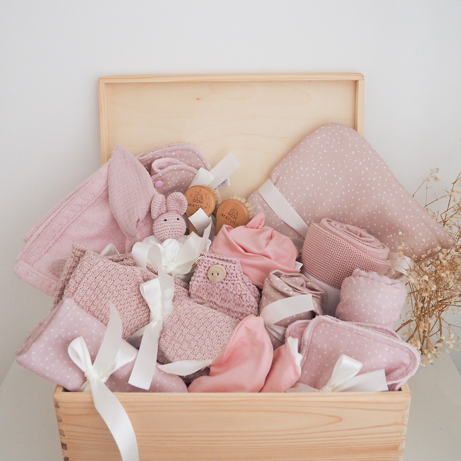 Drewniana skrzynia wypełniona różową wyprawką dla niemowlaka: kocyki w kropki, ręczniki, ubranka i pluszowy króliczek. Całość przewiązana białymi wstążkami.