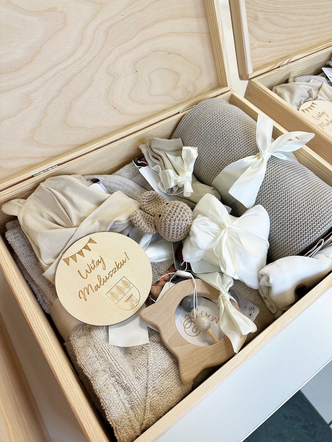 Drewniana skrzynka z prezentami dla noworodka w odcieniach beżu, z drewnianą zabawką i okrągłą ozdobą z napisem 'Witaj Maluszku!'.