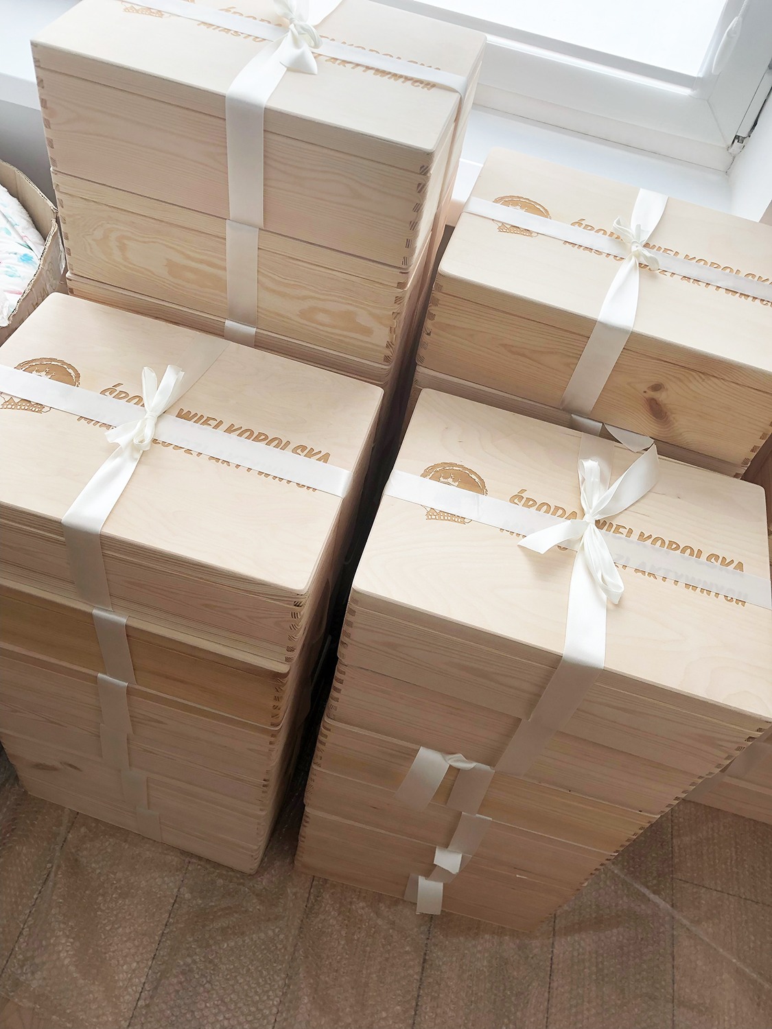 Stosy jasnych, drewnianych skrzynek z logo i białymi wstążkami, gotowych do wręczenia. Widok z góry, naturalne światło.