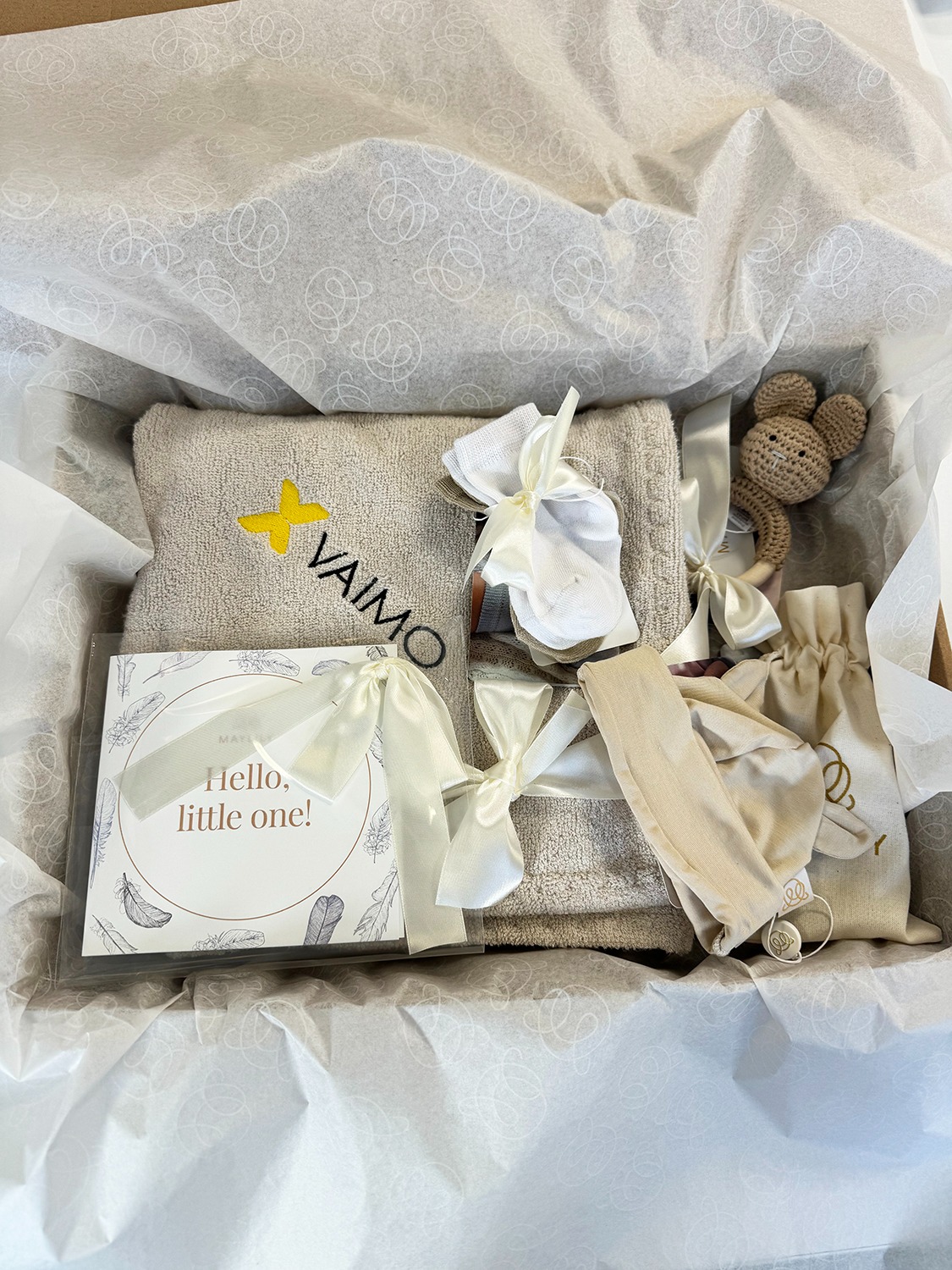 Beżowy ręcznik z logo, gryzak-myszka i kartka powitalna w pudełku prezentowym wyłożonym wzorzystym papierem. Neutralna kolorystyka, idealny upominek dla noworodka.