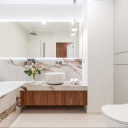 Michał Drożdż Real Estate Broker - Nowoczesna łazienka z marmurowym blatem, umywalką i kwiatami. Widoczna wanna i toaleta. Oświetlenie LED podkreśla elegancję wnętrza.