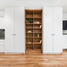 Michał Drożdż Real Estate Broker - Jasne, nowoczesne wnętrze: biała zabudowa kuchenna z drewnianym regałem i telewizorem. Parkiet na podłodze. Minimalistyczny design.