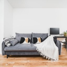 Michał Drożdż Real Estate Broker - Jasne wnętrze: szara sofa z dekoracyjnymi poduszkami i białym pledem, biurko z czarnym blatem i metalowym regałem, drewniana podłoga, duże okno balkonowe.