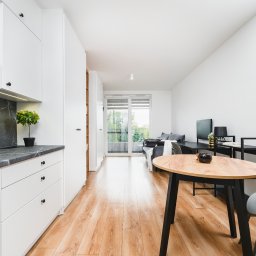 Michał Drożdż Real Estate Broker - Jasne wnętrze mieszkania z drewnianą podłogą, białymi meblami kuchennymi z marmurowym blatem i okrągłym stołem. Widok na balkon i część dzienną. Minimalistyczny design.