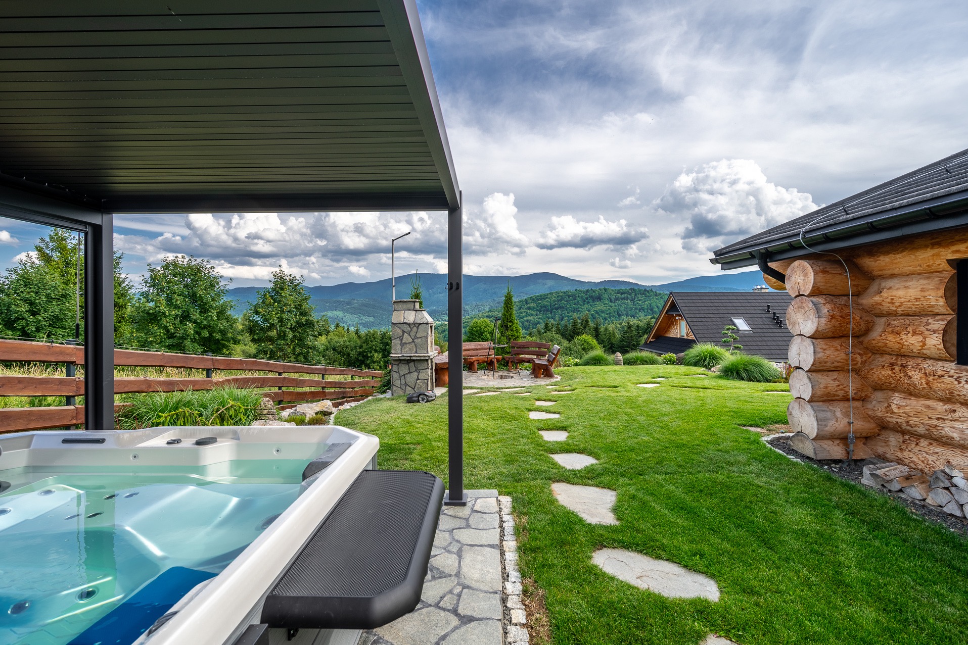 Jacuzzi na tarasie drewnianego domu z widokiem na góry. Kamienne ścieżki prowadzą przez trawnik do paleniska z ławkami. Nowoczesny design i relaks w otoczeniu natury.