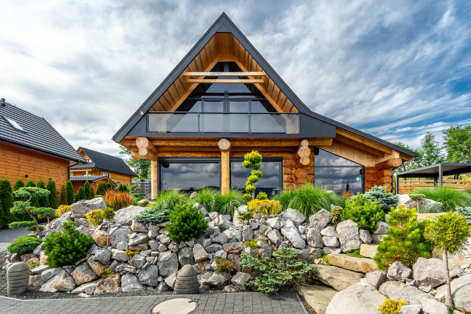 Nowoczesny dom z bali z dużymi oknami i kamiennym ogrodem. Drewniana elewacja, czarny dach, minimalistyczny design. W tle błękitne niebo z chmurami.