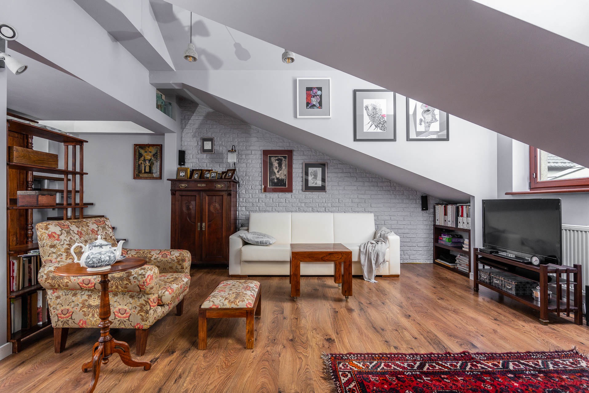 Jasne poddasze ze skośnym sufitem i ceglaną ścianą. Wnętrze urządzone w stylu retro z drewnianą podłogą, sofą, fotelem i dywanem. Widoczne meble i dekoracje.