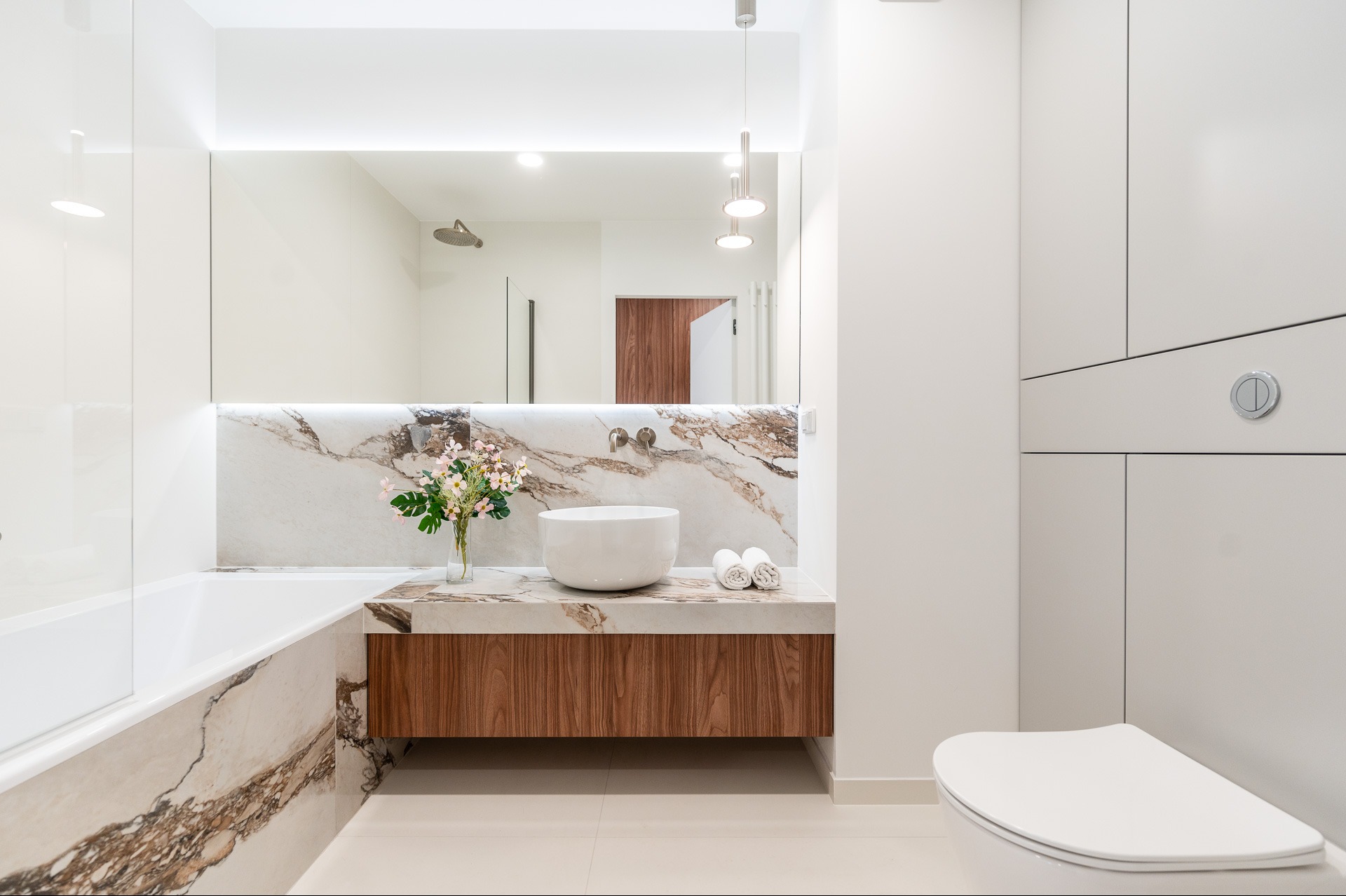 Nowoczesna łazienka z marmurowym blatem, umywalką i kwiatami. Widoczna wanna i toaleta. Oświetlenie LED podkreśla elegancję wnętrza.