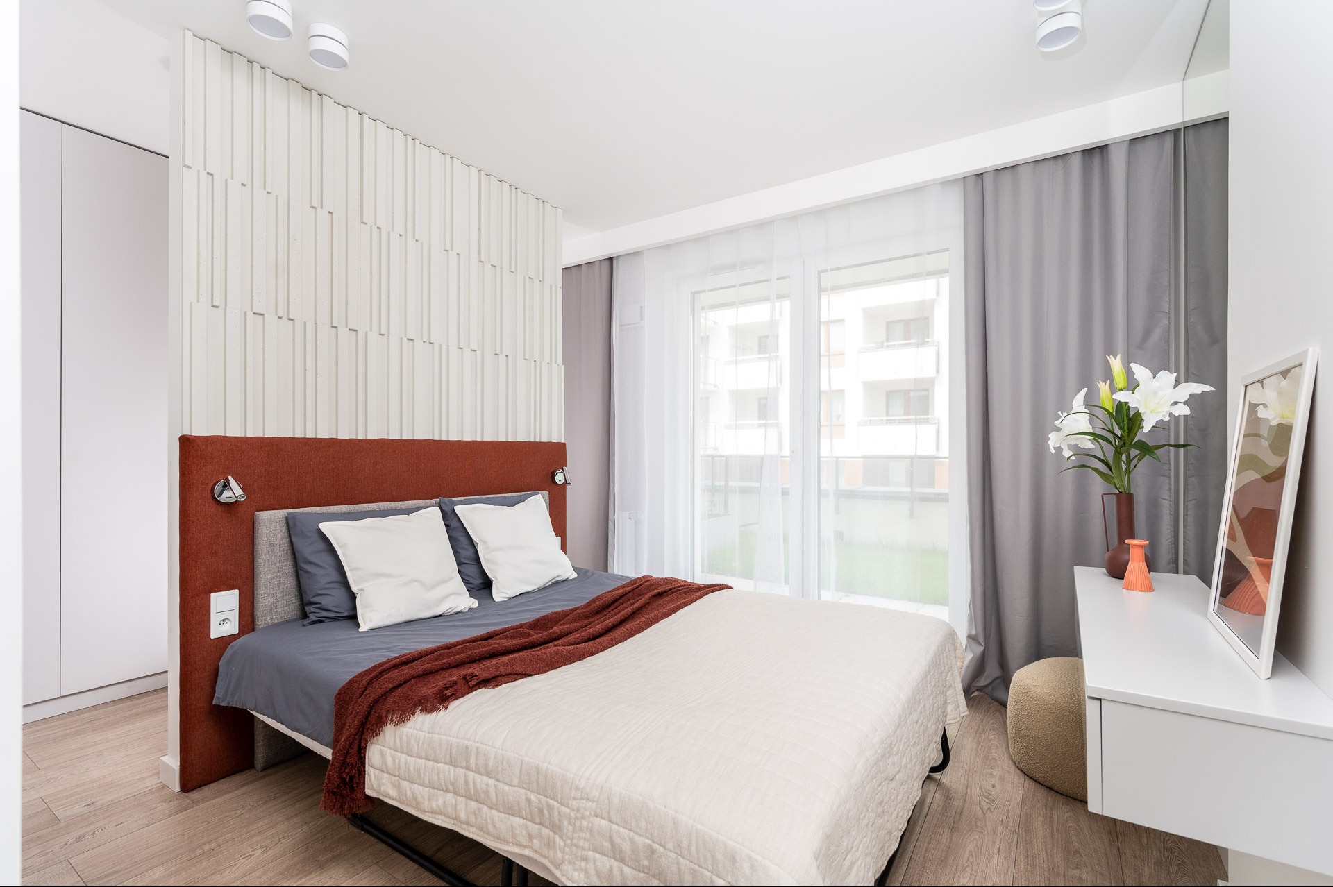 Nowoczesna sypialnia z łóżkiem, stolikiem i dekoracjami. Stonowana kolorystyka, drewniana podłoga. Widok na budynek przez okno z zasłonami.