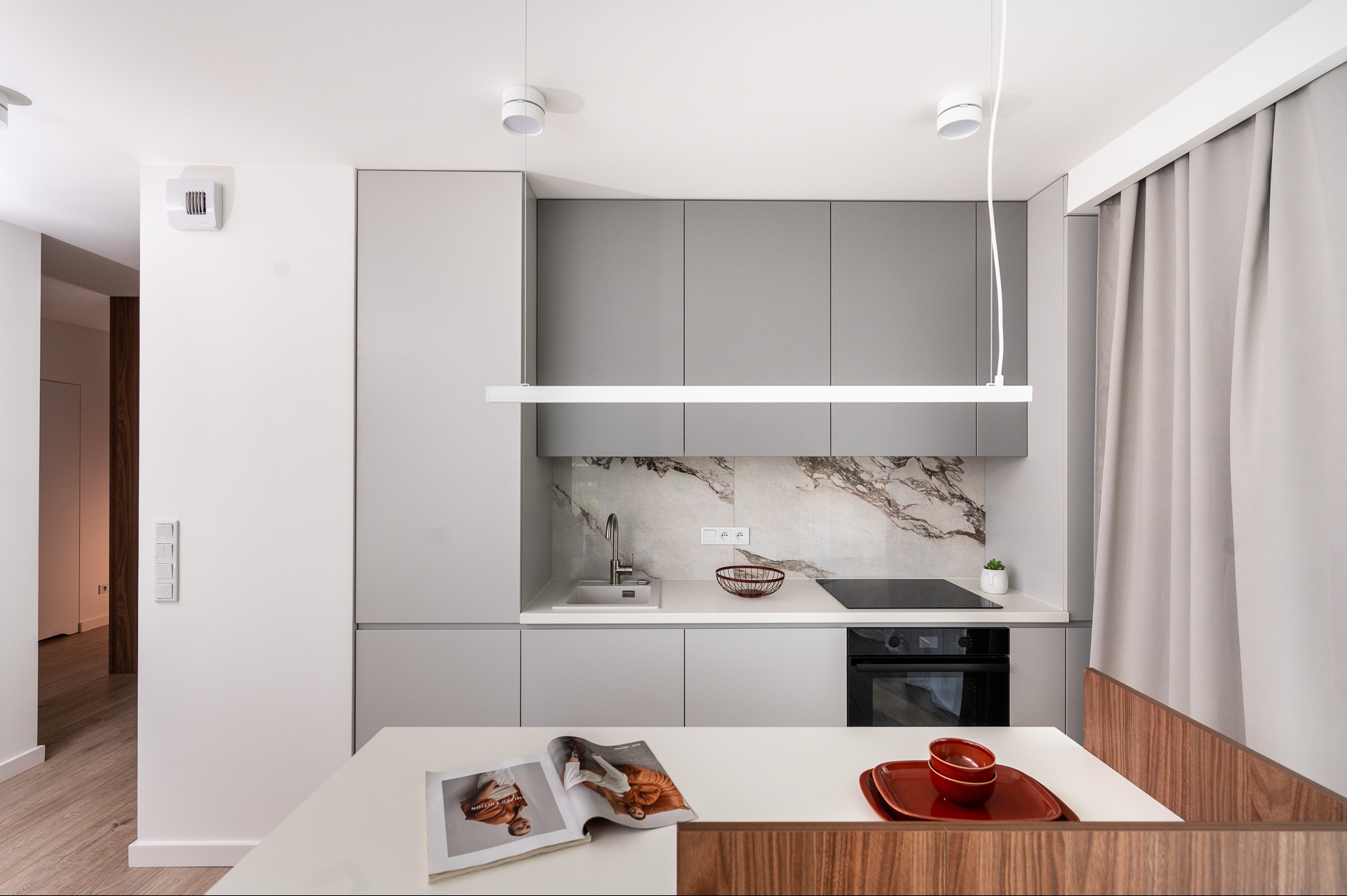 Nowoczesna, minimalistyczna kuchnia w odcieniach szarości z marmurowym backsplash, oświetleniem LED i drewnianymi akcentami przy stole. Widok na magazyn na blacie.