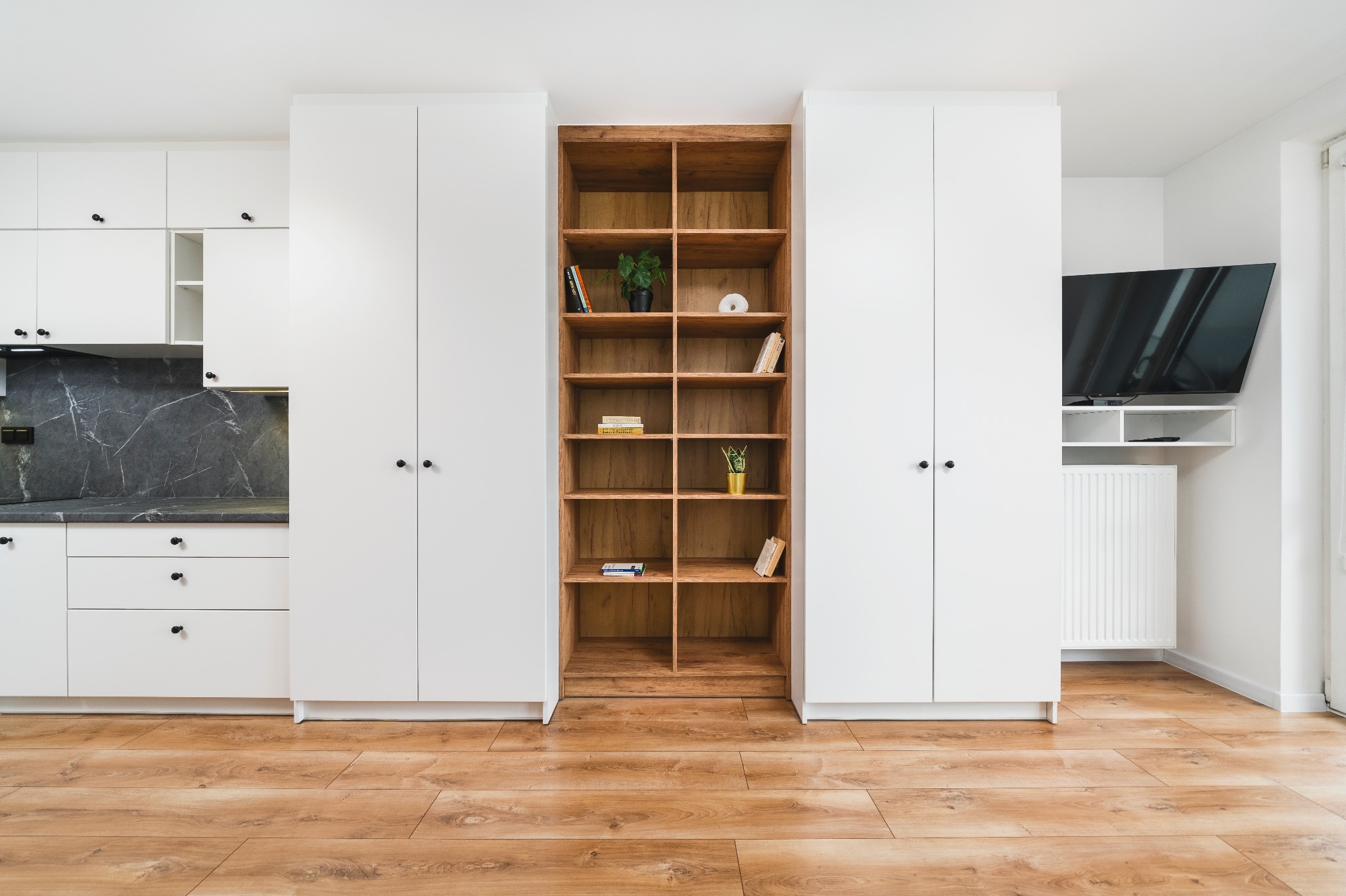 Jasne, nowoczesne wnętrze: biała zabudowa kuchenna z drewnianym regałem i telewizorem. Parkiet na podłodze. Minimalistyczny design.