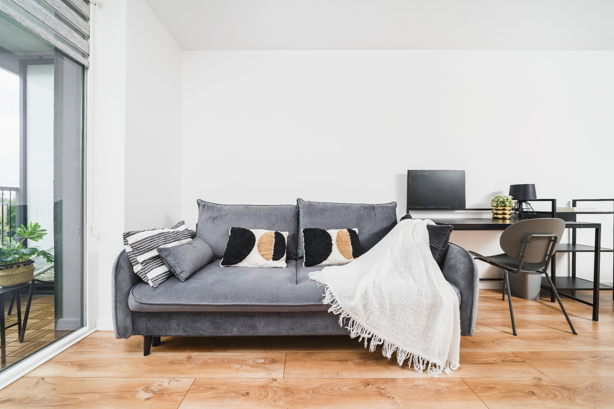 Jasne wnętrze: szara sofa z dekoracyjnymi poduszkami i białym pledem, biurko z czarnym blatem i metalowym regałem, drewniana podłoga, duże okno balkonowe.