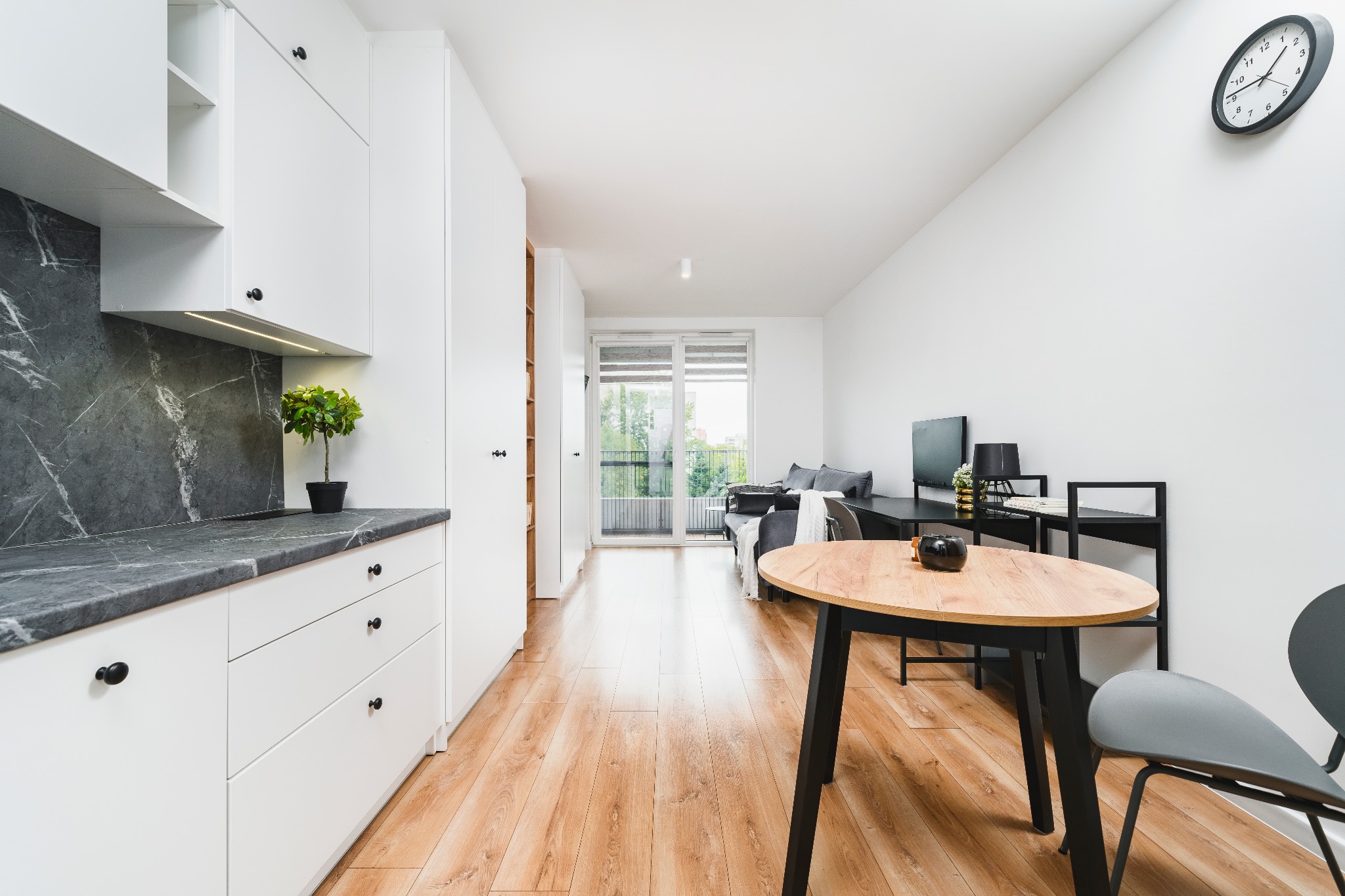 Jasne wnętrze mieszkania z drewnianą podłogą, białymi meblami kuchennymi z marmurowym blatem i okrągłym stołem. Widok na balkon i część dzienną. Minimalistyczny design.