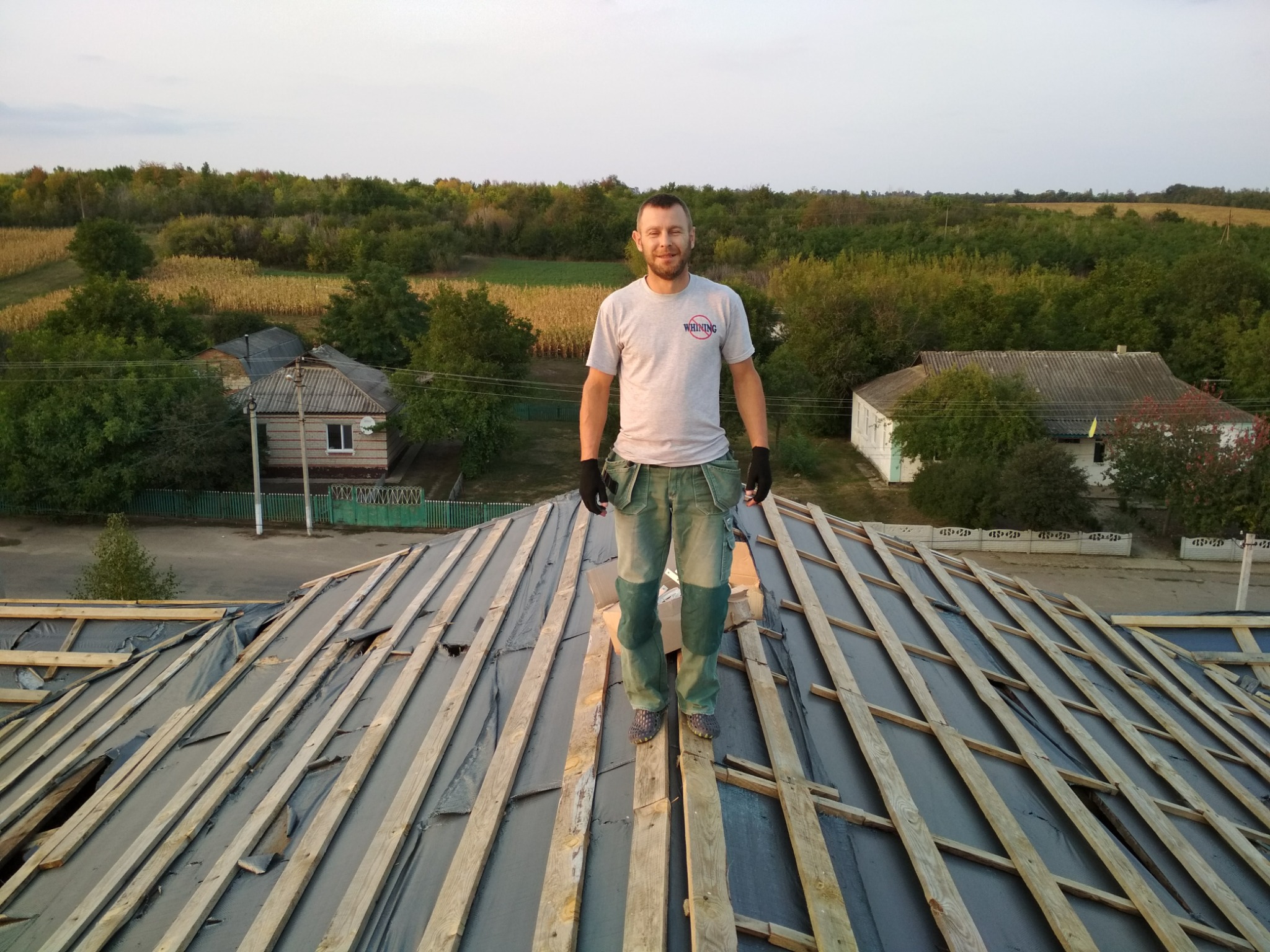 Mężczyzna w roboczym stroju stoi na drewnianej konstrukcji dachu domu jednorodzinnego na tle zieleni i nieba. Widoczne deski i czarna membrana dachowa.