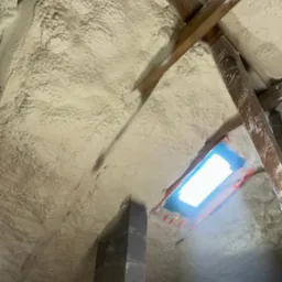 Poddasze po natrysku pianki PUR, widoczne drewniane elementy konstrukcyjne i okno dachowe zabezpieczone folią. Izolacja pokrywa całą powierzchnię.