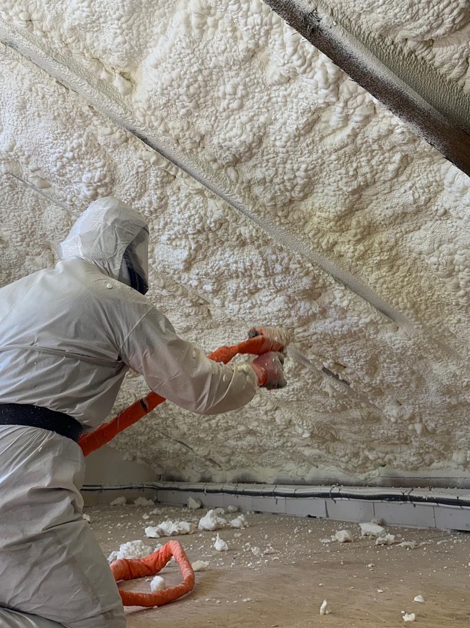 Aplikacja pianki PUR na poddaszu przez pracownika w kombinezonie ochronnym, widoczny wąż i świeżo nałożona, spieniona izolacja. Detal: widoczne przewody elektryczne.