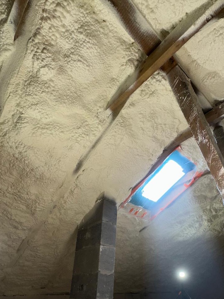 Poddasze po natrysku pianki PUR, widoczne drewniane elementy konstrukcyjne i okno dachowe zabezpieczone folią. Izolacja pokrywa całą powierzchnię.