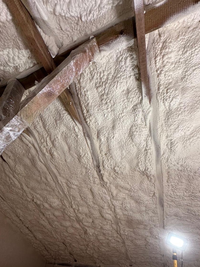 Poddasze po ociepleniu pianką PUR, widoczne drewniane elementy konstrukcji. Pianka o nierównej fakturze, szczelnie wypełnia przestrzeń. Narożnik oświetlony lampą.