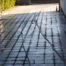 Mokry chodnik z kostki brukowej po czyszczeniu, wąż ciśnieniowy na powierzchni, widoczne ślady wody i różnica w kolorze kostki, budynek i zieleń w tle.