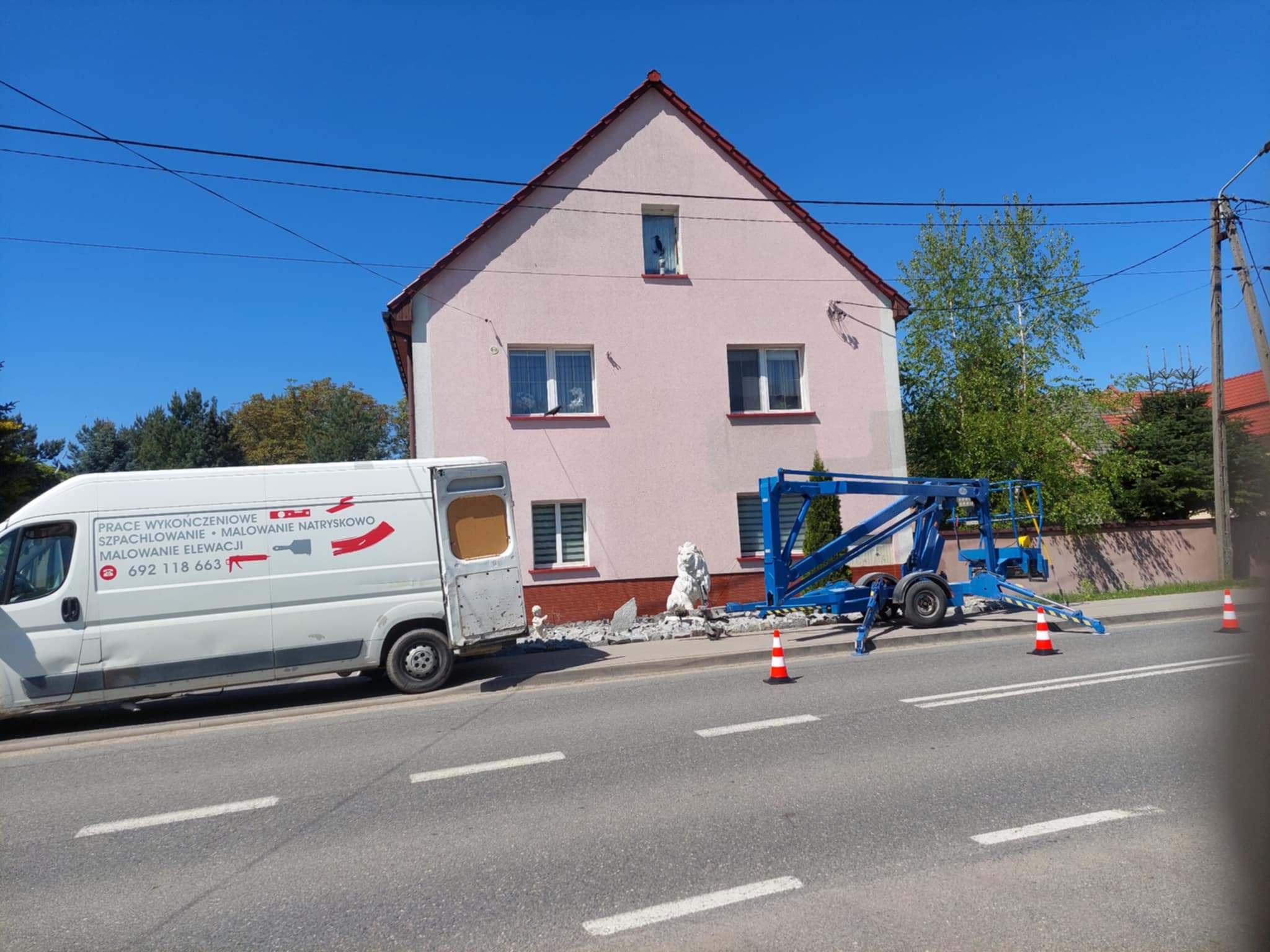 Różowy dom z elewacją w trakcie naprawy, widoczny podnośnik koszowy i gruz. Na pierwszym planie samochód z napisem o pracach wykończeniowych i malowaniu natryskowym elewacji.