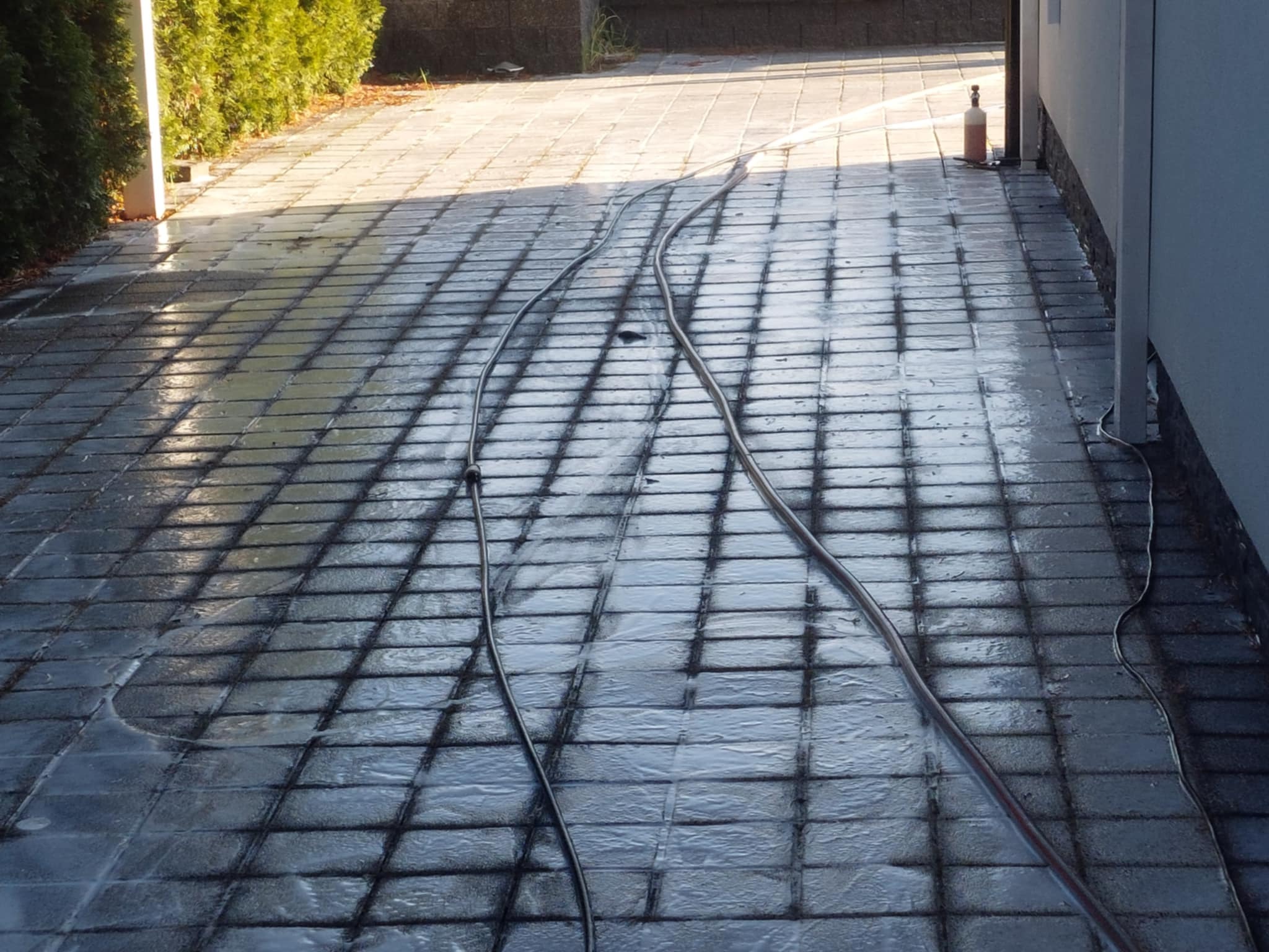 Mokry chodnik z kostki brukowej po czyszczeniu, wąż ciśnieniowy na powierzchni, widoczne ślady wody i różnica w kolorze kostki, budynek i zieleń w tle.