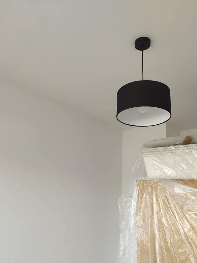 Czarna lampa sufitowa z białym wnętrzem klosza w jasnym wnętrzu. Meble zabezpieczone folią malarską. Minimalistyczny design oświetlenia.
