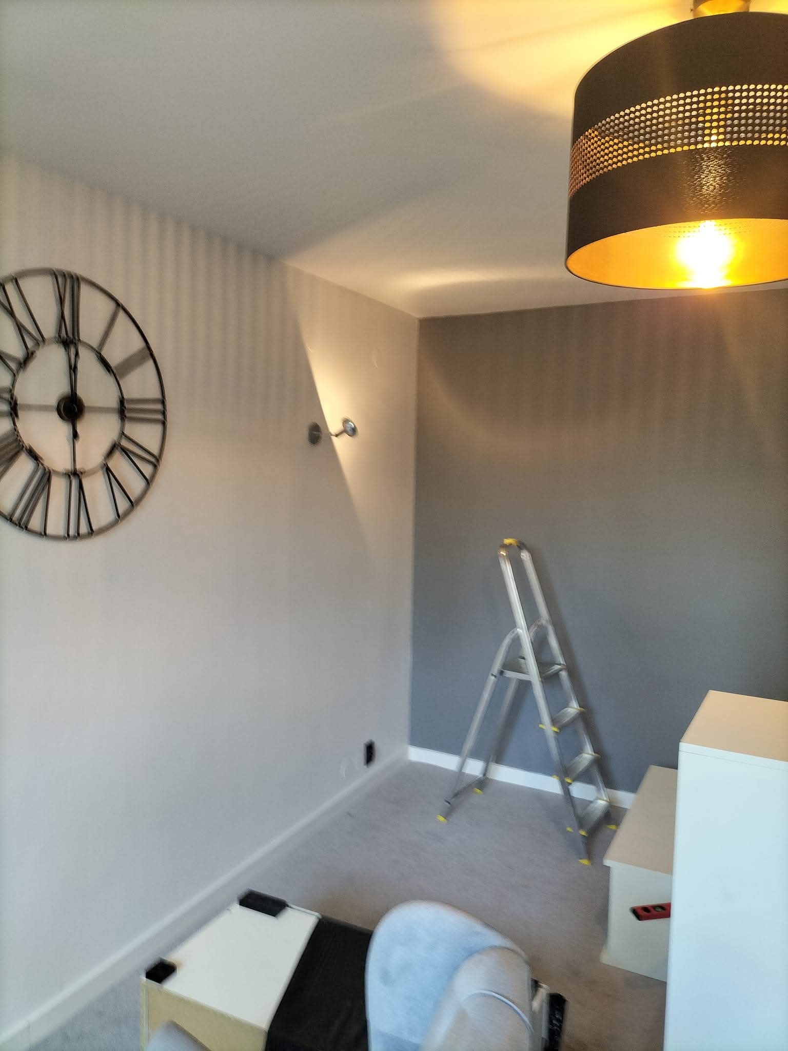 Świeżo pomalowane wnętrze pokoju w odcieniach szarości z dekoracyjnym zegarem na ścianie i drabiną opartą o ścianę. Widoczna lampa sufitowa i meble.