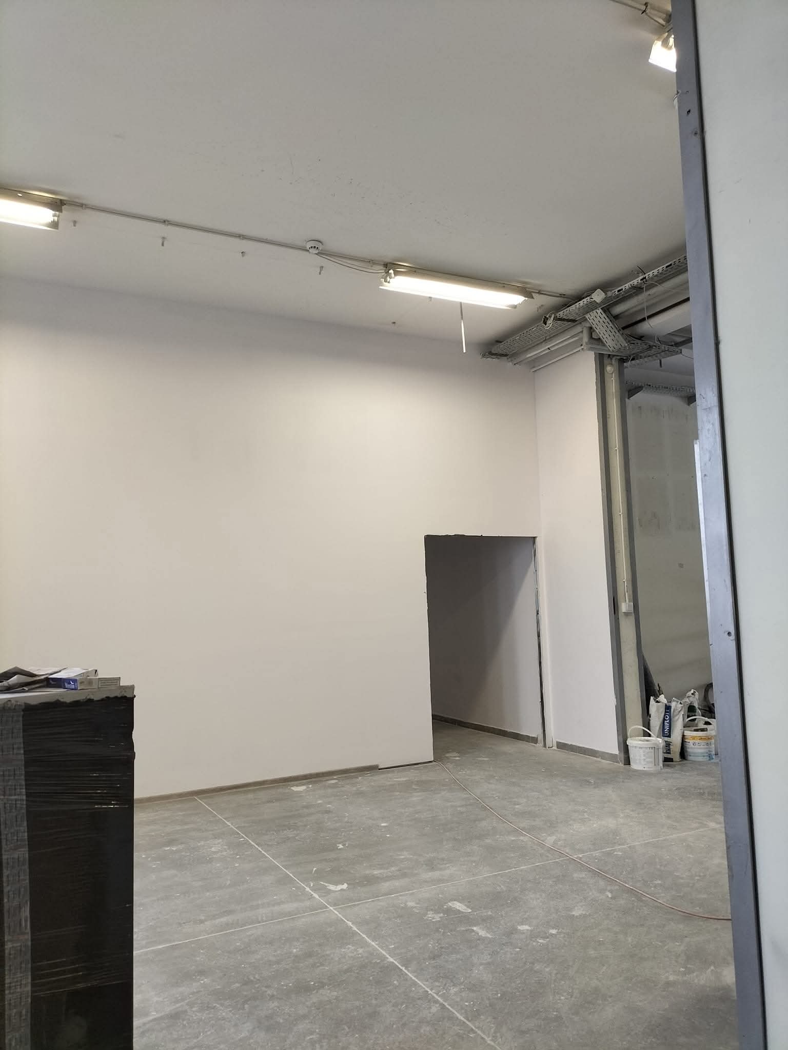 Wnętrze po szpachlowaniu i malowaniu ścian na biało, widoczne puszki z farbą, surowa podłoga, instalacje pod sufitem i otwór drzwiowy.