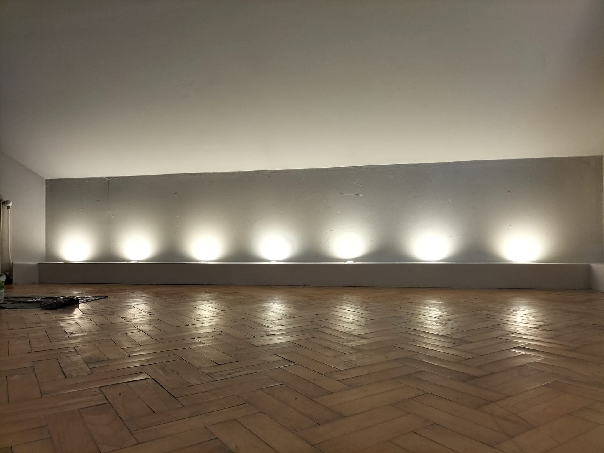 Parkiet w jodełkę, oświetlona ściana z wnęką i punktowymi lampami. Widoczne narzędzia malarskie w rogu, sugerujące świeżo zakończone prace wykończeniowe.