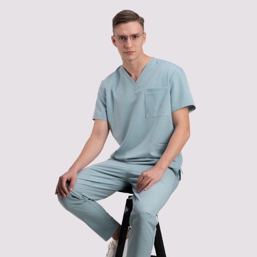 Mężczyzna w okularach, siedzący na stołku, prezentuje kompletny, błękitny strój medyczny z krótkim rękawem i kieszenią na bluzie, oraz dopasowane spodnie.