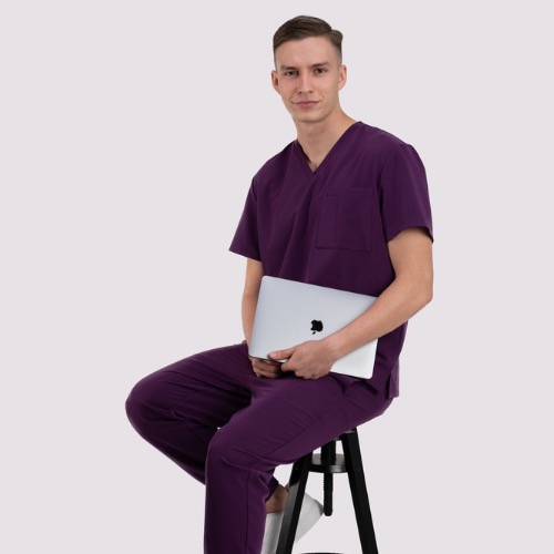 Mężczyzna siedzi na stołku, ubrany w śliwkowy uniform medyczny, trzymając laptopa. Stonowane tło, oświetlenie studyjne, elegancki krój uniformu.