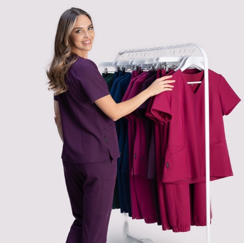 Uśmiechnięta kobieta prezentuje kolekcję kolorowych, damskich uniformów medycznych na wieszaku, w tym fioletowy komplet, na jasnym tle.