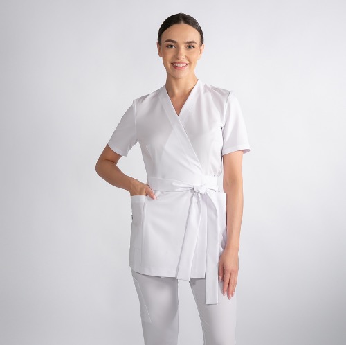 Uśmiechnięta kobieta prezentuje białą tunikę medyczną z wiązaniem w pasie i spodnie, na jasnym tle. Strój przeznaczony dla personelu medycznego.