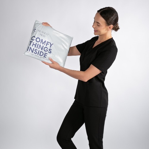 Uśmiechnięta kobieta w czarnym uniformie medycznym prezentuje zapakowane zamówienie Blanco Med z napisem 'Comfy things inside' na jasnoszarym tle.