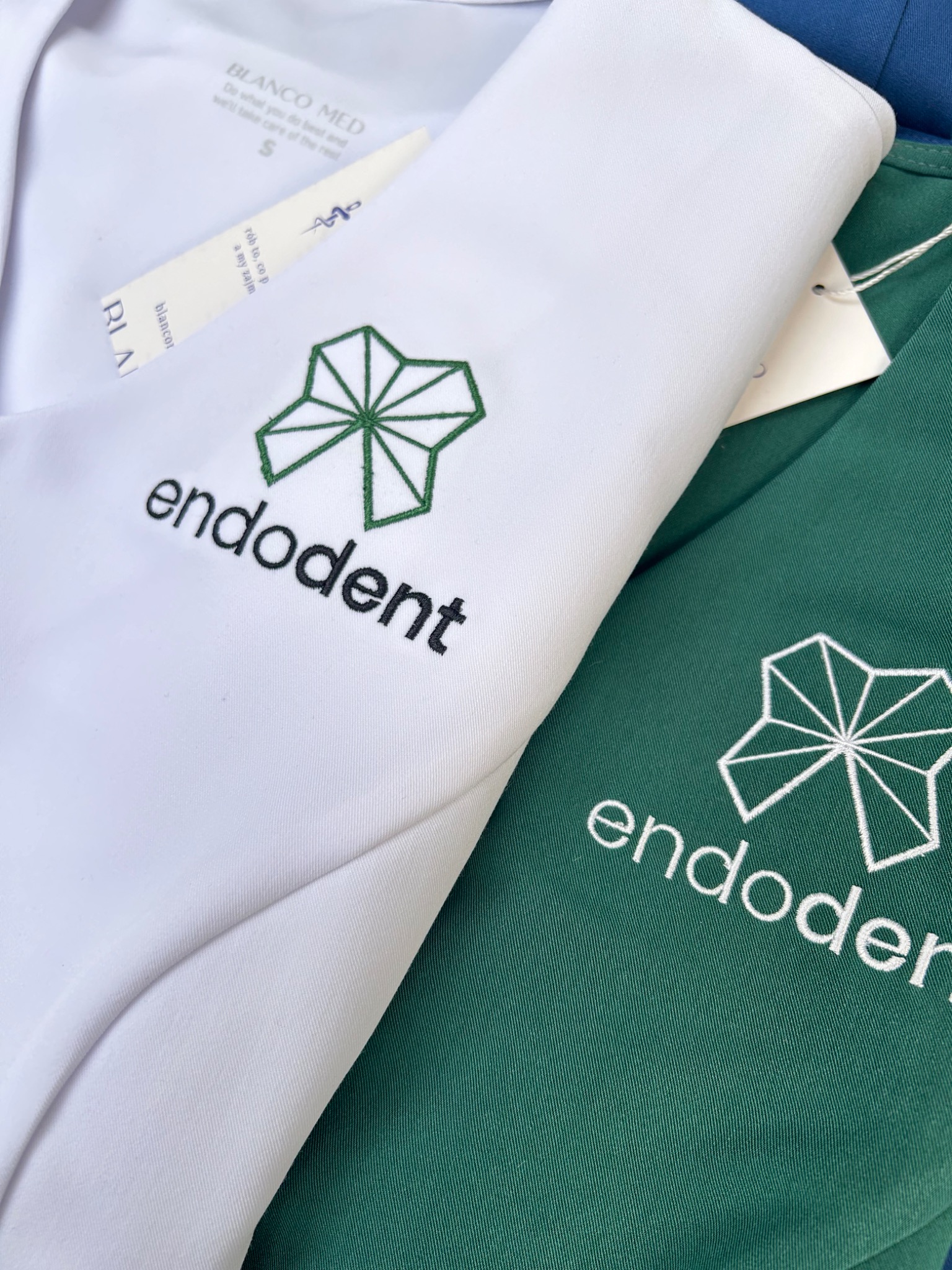 Dwa uniformy medyczne, biały i zielony, z wyhaftowanym logo i napisem 'endodent'. Widoczna metka Blanco Med. Ubrania schludnie ułożone, prezentacja detali.