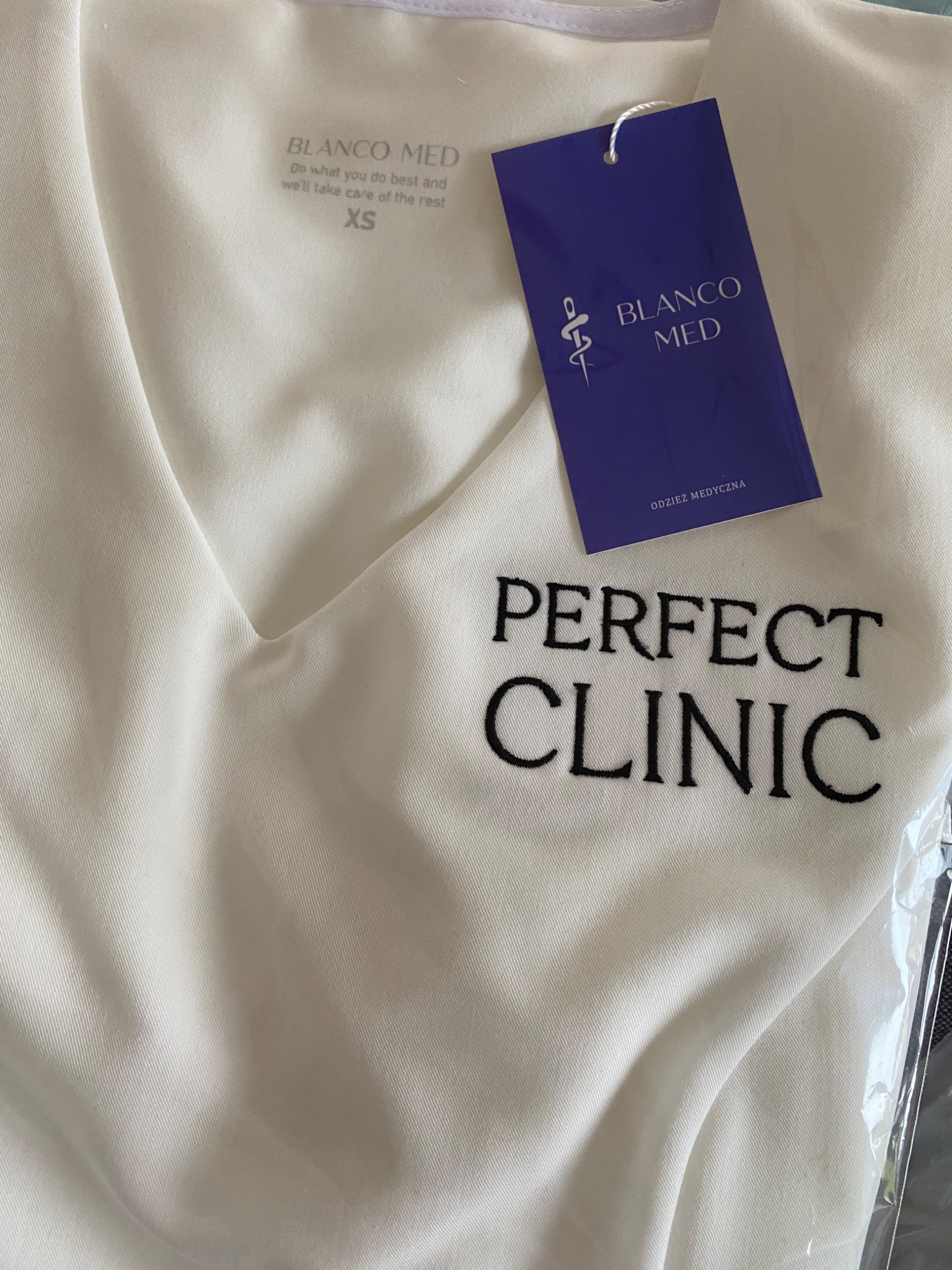 Biała bluza medyczna z haftowanym logo 'Perfect Clinic' i metką 'Blanco Med' w zbliżeniu, prezentująca odzież dla personelu medycznego.