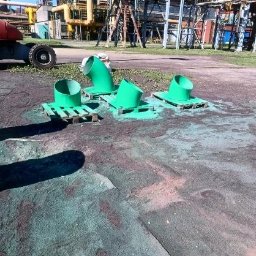 TOPREM SPÓŁKA Z OGRANICZONĄ ODPOWIEDZIALNOŚCIĄ - Cztery zielone rury na paletach po malowaniu, na tle instalacji przemysłowej. Widoczne ślady farby na podłożu, co sugeruje niedawne malowanie.