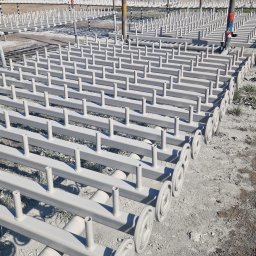 TOPREM SPÓŁKA Z OGRANICZONĄ ODPOWIEDZIALNOŚCIĄ - Metalowe konstrukcje po obróbce, pokryte jednolitą szarą farbą, ułożone rzędami na zewnątrz, z widoczną infrastrukturą przemysłową w tle.