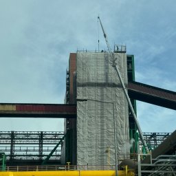 TOPREM SPÓŁKA Z OGRANICZONĄ ODPOWIEDZIALNOŚCIĄ - Przemysłowy budynek w trakcie prac renowacyjnych, osłonięty siatką, z dźwigiem i widocznymi rurociągami. Elementy konstrukcyjne w kolorach zielonym i żółtym.