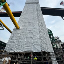 TOPREM SPÓŁKA Z OGRANICZONĄ ODPOWIEDZIALNOŚCIĄ - Wysoka konstrukcja przemysłowa osłonięta siatką ochronną na rusztowaniu, widoczny pracownik w stroju roboczym, fragmenty instalacji i kominów w tle.